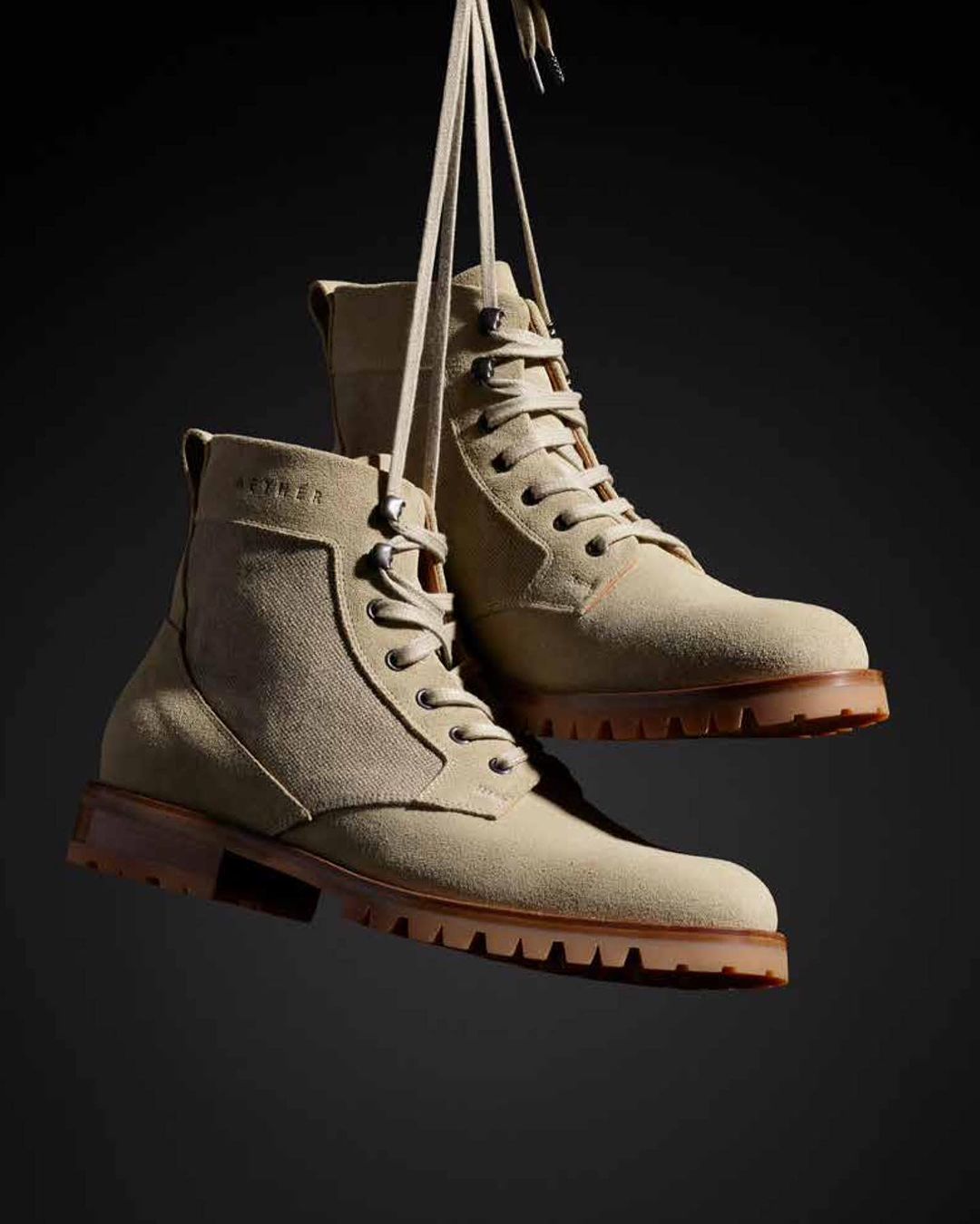 Beige suede ankle boots hanging by their laces against a dark backdrop in a studio shot, capturing their textured material and design.