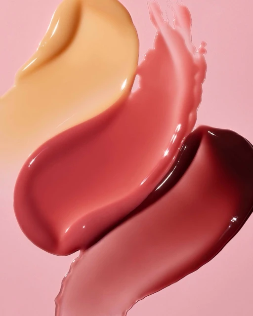 Close-up of swatches of lip gloss in varying shades of pink, red, and nude against a soft pink background, highlighting texture.