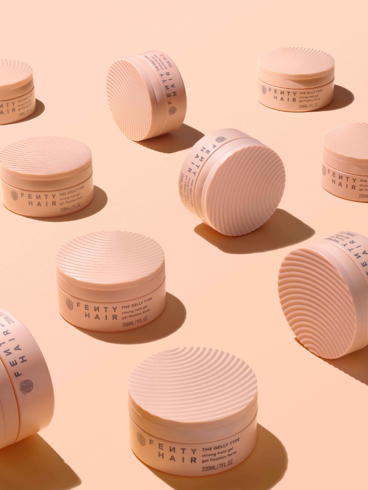 Several containers of Fenty Hair The Belly Type strong hold gel for edges are arranged on a peach-colored surface in a studio shot.