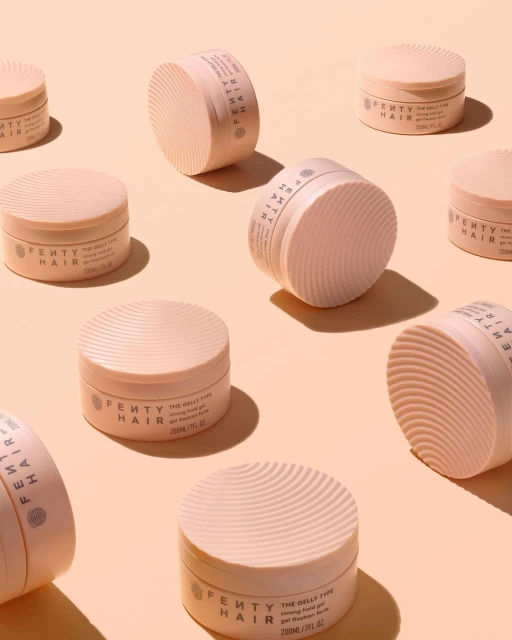 Multiple pink Fenty Hair gel products arranged on a light peach background in studio lighting, beauty shot for product marketing.