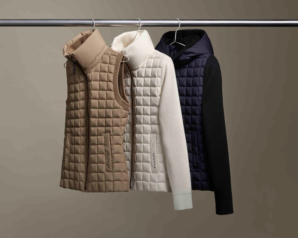 Three quilted vests and a jacket hang on a silver metal clothing rack against a neutral beige background in a studio setting.