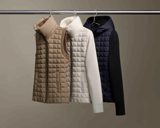 Three quilted vests and a jacket hang on a silver metal clothing rack against a neutral beige background in a studio setting.