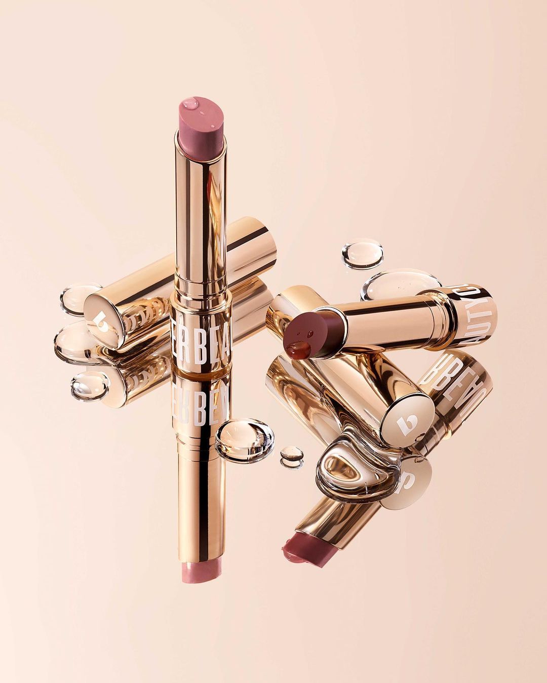 Close-up studio shot of a variety of lipstick tubes with gold cases and water droplets on a reflective light pink surface.