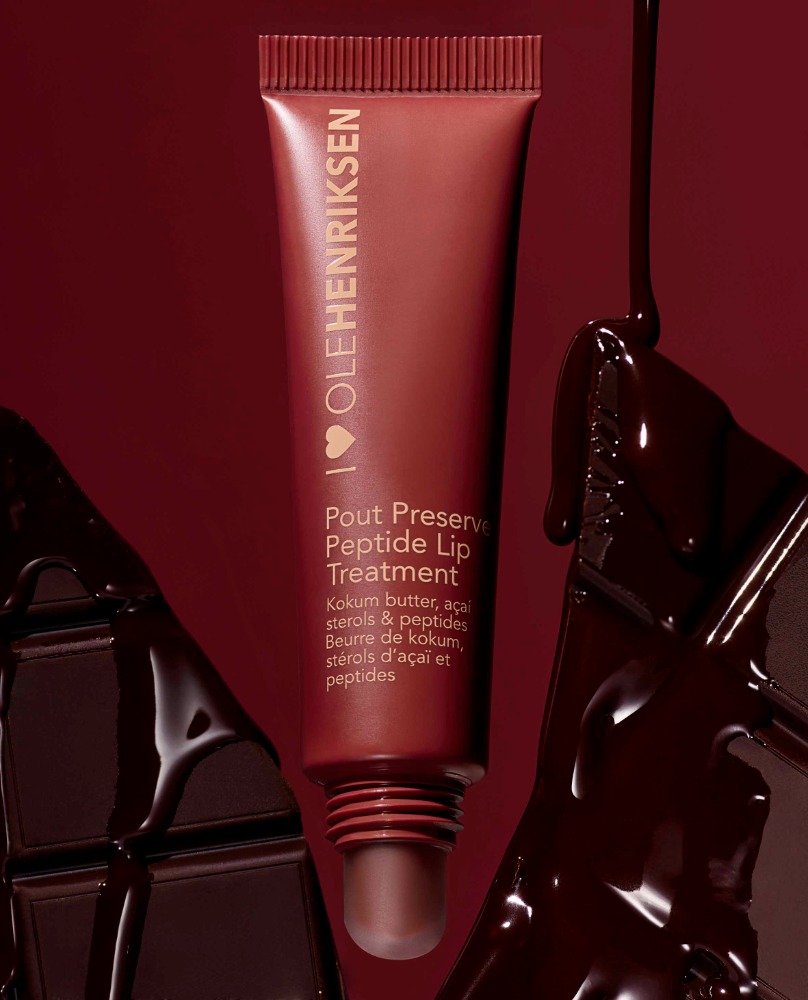 Olehenriksen Pout Preserve Peptide Lip Treatment tube with dark chocolate chunks on a dark red background in a studio shot.