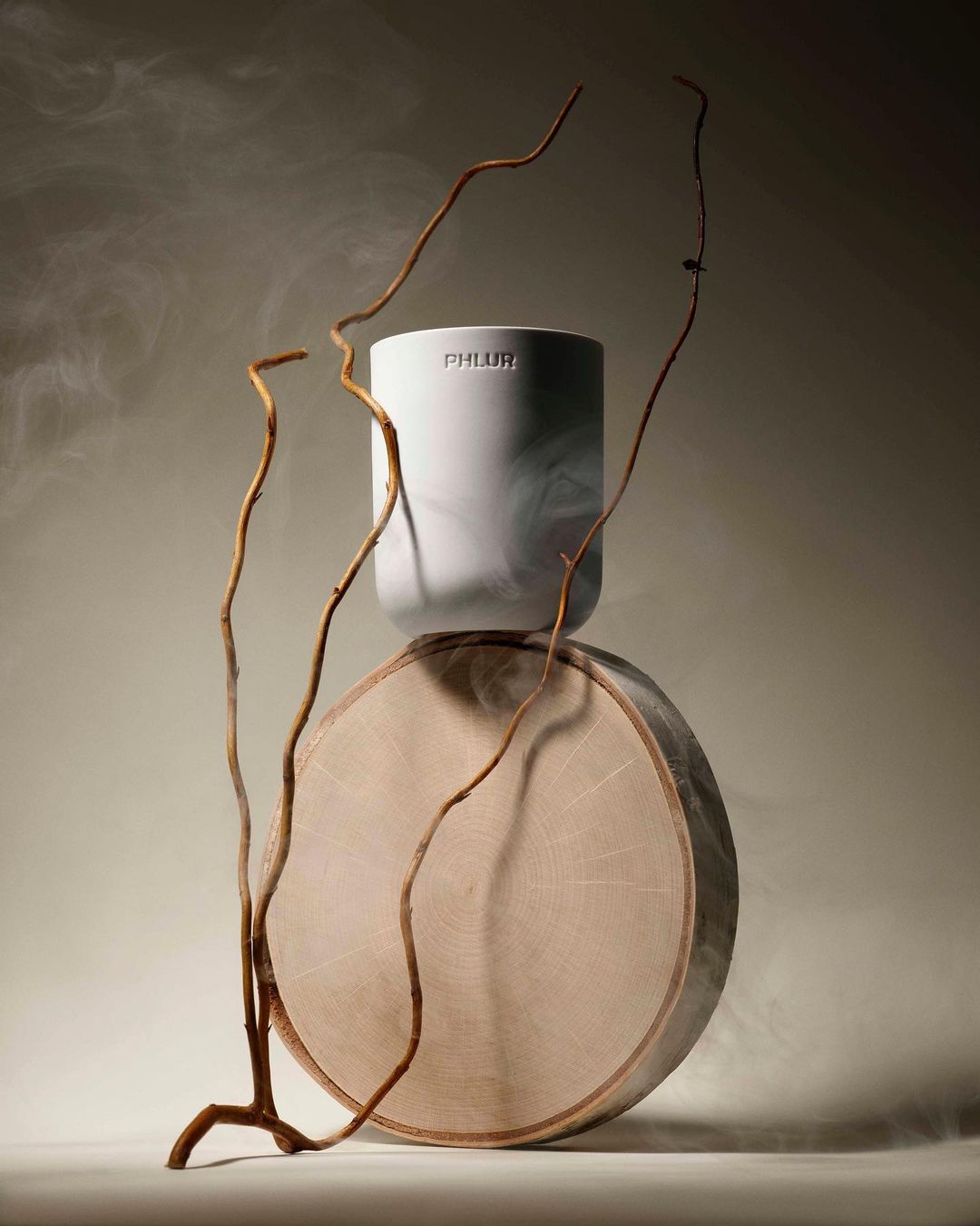 Still life featuring a white Phlur candle on a round wooden base, accented with bare branches against a smoky beige background.