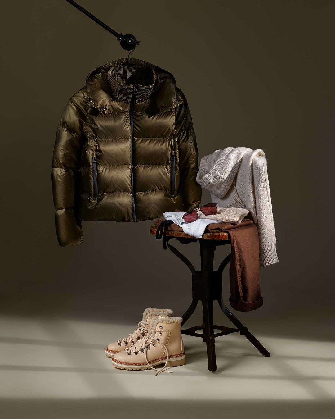 Padded jacket on hanger with boots, sunglasses, and clothing on stool against neutral background, capturing a stylish fashion still life.