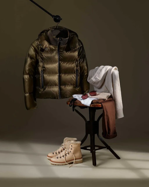 Padded jacket on hanger with boots, sunglasses, and clothing on stool against neutral background, capturing a stylish fashion still life.