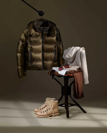 Padded jacket on hanger with boots, sunglasses, and clothing on stool against neutral background, capturing a stylish fashion still life.
