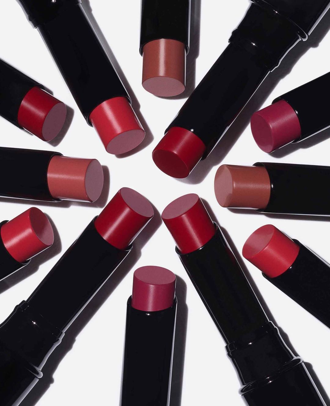 Various shades of lipstick in black tubes are arranged in a circular pattern on a white background. Beauty and cosmetic product photography.