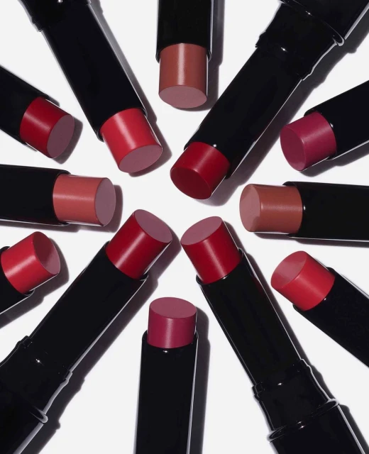 Various shades of lipstick in black tubes are arranged in a circular pattern on a white background. Beauty and cosmetic product photography.