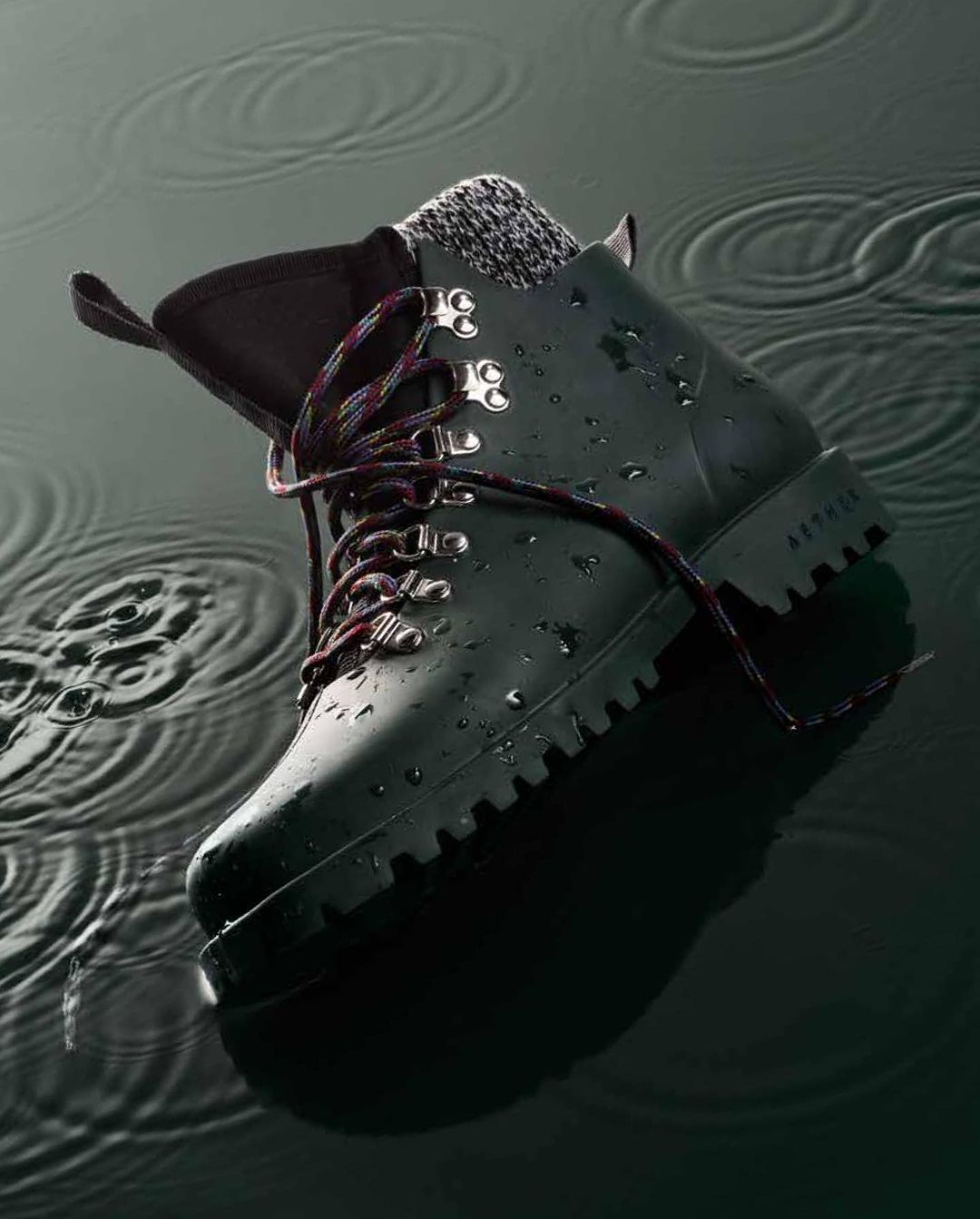 Elevated studio shot of a green Aether hiking boot with gray sock and colorful laces resting in a dark liquid with ripple patterns.