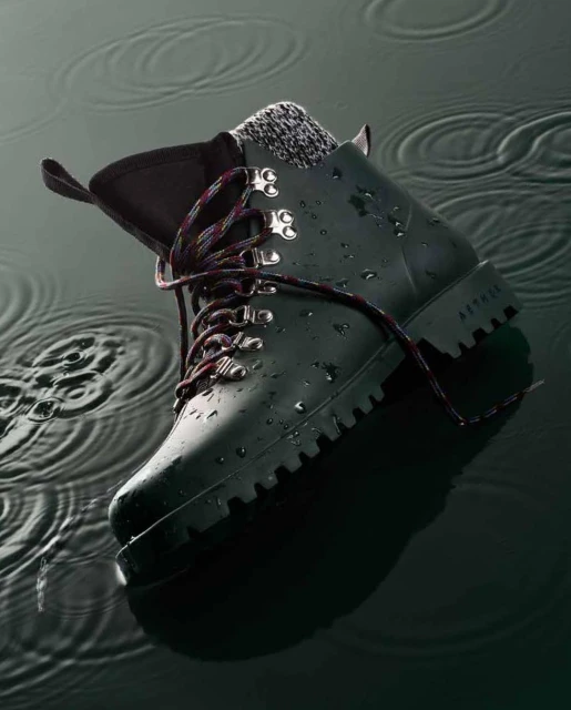 Elevated studio shot of a green Aether hiking boot with gray sock and colorful laces resting in a dark liquid with ripple patterns.