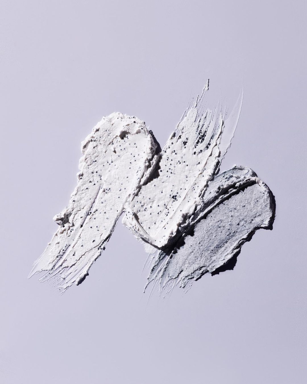 Three swipes of cream with scrub granules on a light purple surface. Cosmetic product shot for beauty, skincare, or health.