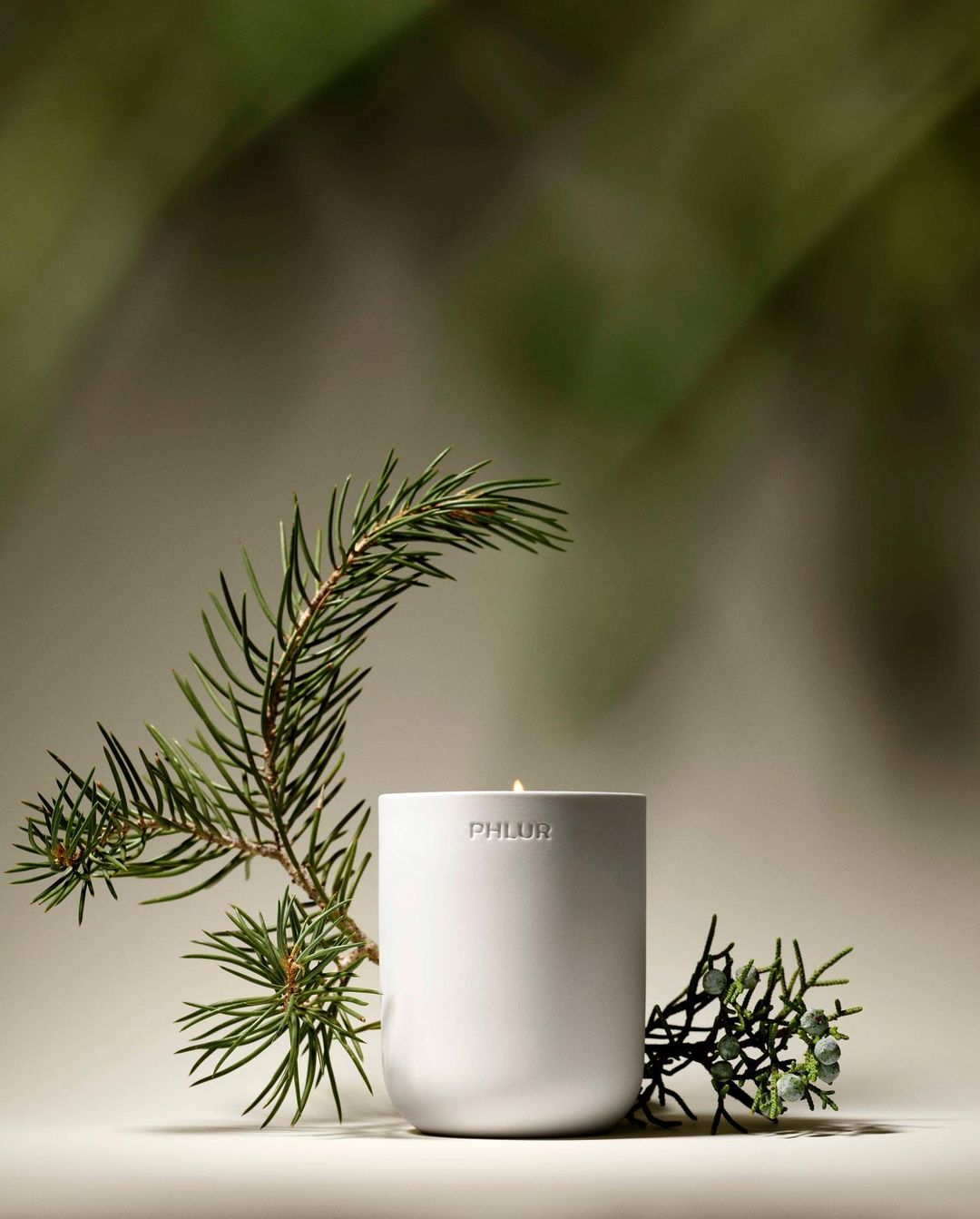 White candle with Phlur logo on a beige surface next to green pine branches against a blurred green background.