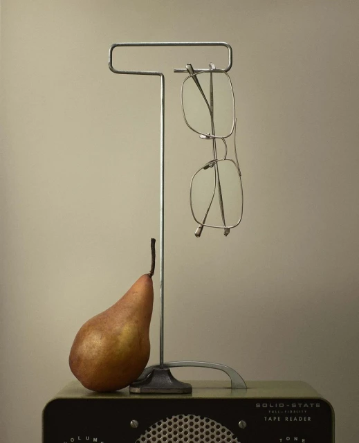 Still life of a pear resting on a vintage radio next to a metal glasses stand with eyeglasses, against a neutral-colored background.