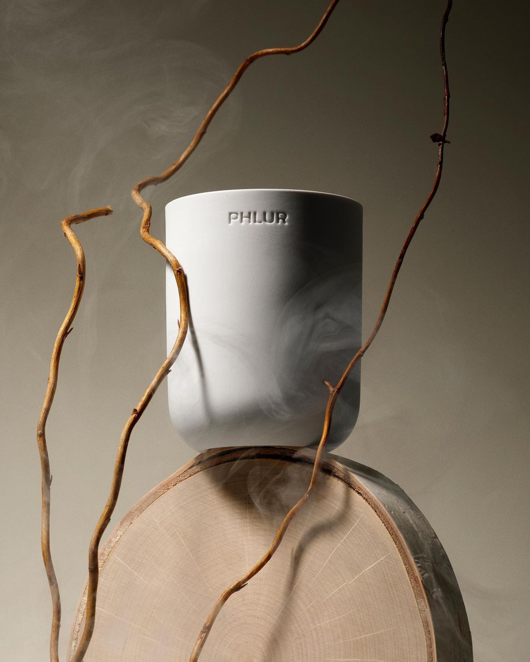 Still life of a PHLUR brand container on a wood base, surrounded by branches and smoke against a tan backdrop. Product shot.