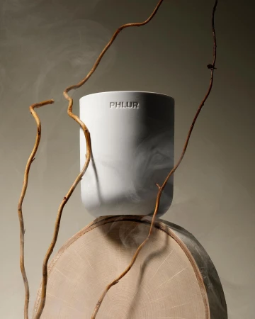 White ceramic PHLUR candle vessel on a wooden slice prop with thin dried branches and wisps of smoke against a neutral background.