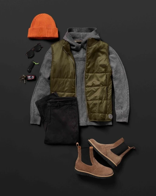 Flat lay of menswear including a gray sweater, olive vest, black pants, brown suede boots, orange beanie, and sunglasses.