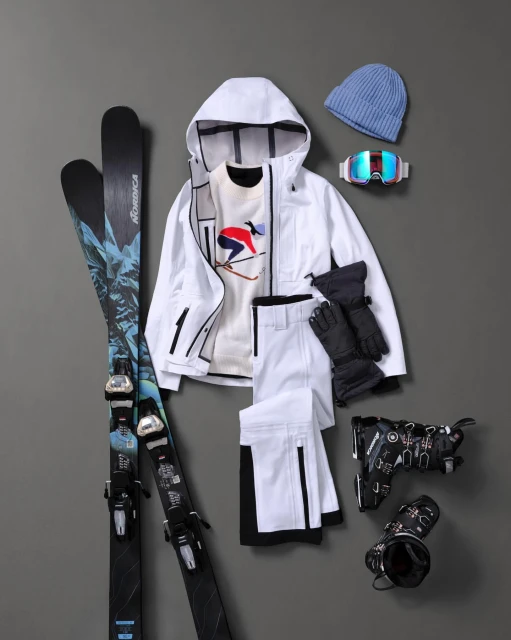 Ski apparel and equipment: a white hooded jacket, ski pants, gloves, hat, goggles, skis, and boots are styled on a gray surface.