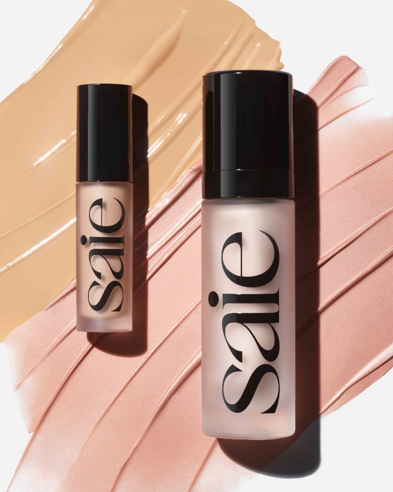 Two Saie cosmetic bottles placed on top of beige and pink liquid makeup swatches against a plain white background.