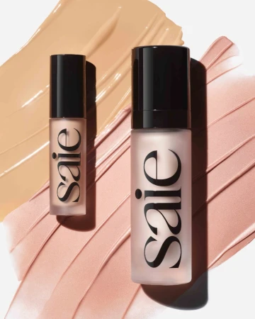 Two Saie cosmetic bottles placed on top of beige and pink liquid makeup swatches against a plain white background.