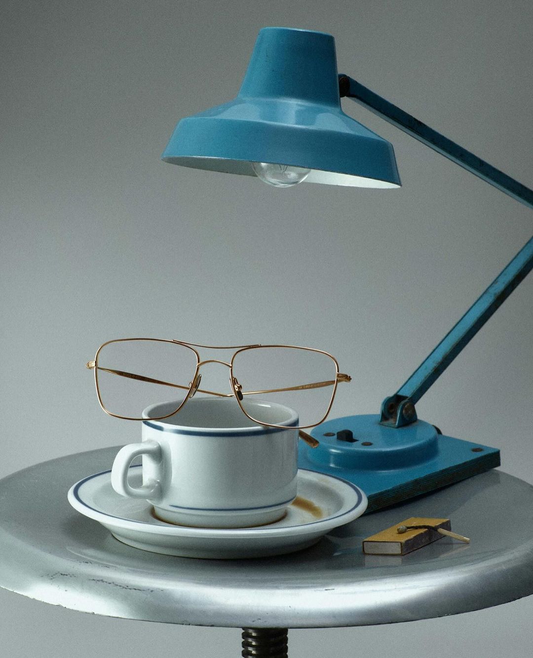 Still life shot features a blue desk lamp, eyeglasses on a white coffee cup, saucer, matchbox on a small metal table with a gray background.
