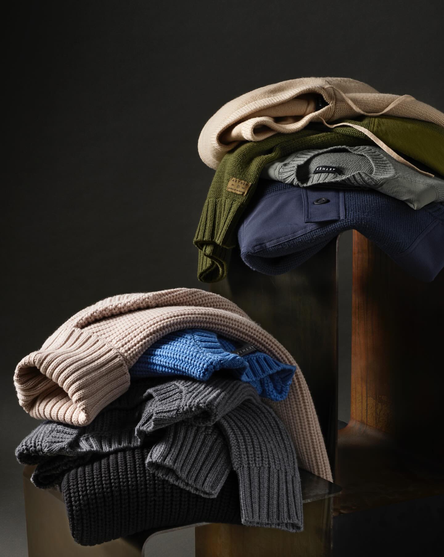 Stacked sweaters and shirts in neutral tones on a reflective surface against a dark background. Fashion and style apparel product shot.