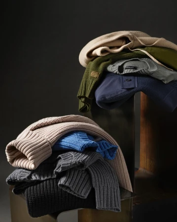 Stacked sweaters and shirts in neutral tones on a reflective surface against a dark background. Fashion and style apparel product shot.