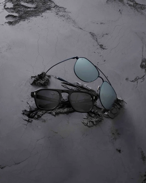 Two pairs of black sunglasses laying on a textured grey surface, one with aviator-style lenses and one with rectangular frames.