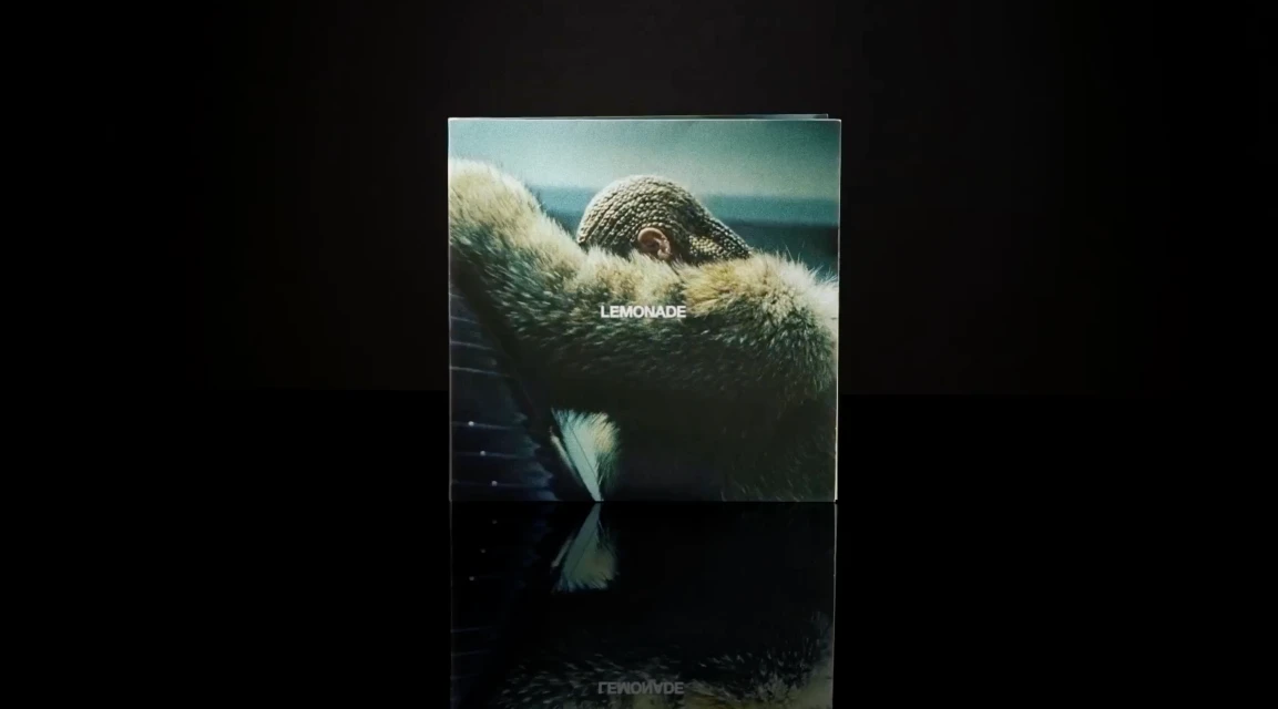 Beyoncé's 'Lemonade' album cover, features the singer with cornrow braids and wearing a fur coat, displayed on a reflective surface.