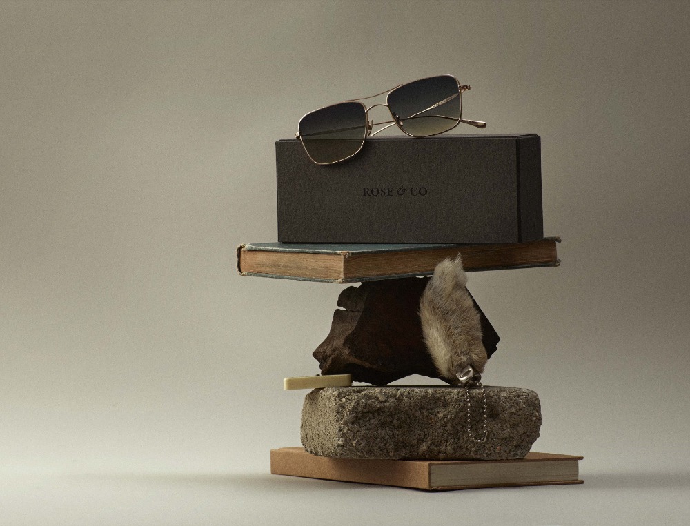 Still life photo of sunglasses and a box on a stacked stone, books, and wood block against a gray background. Fashion and accessory aesthetic.
