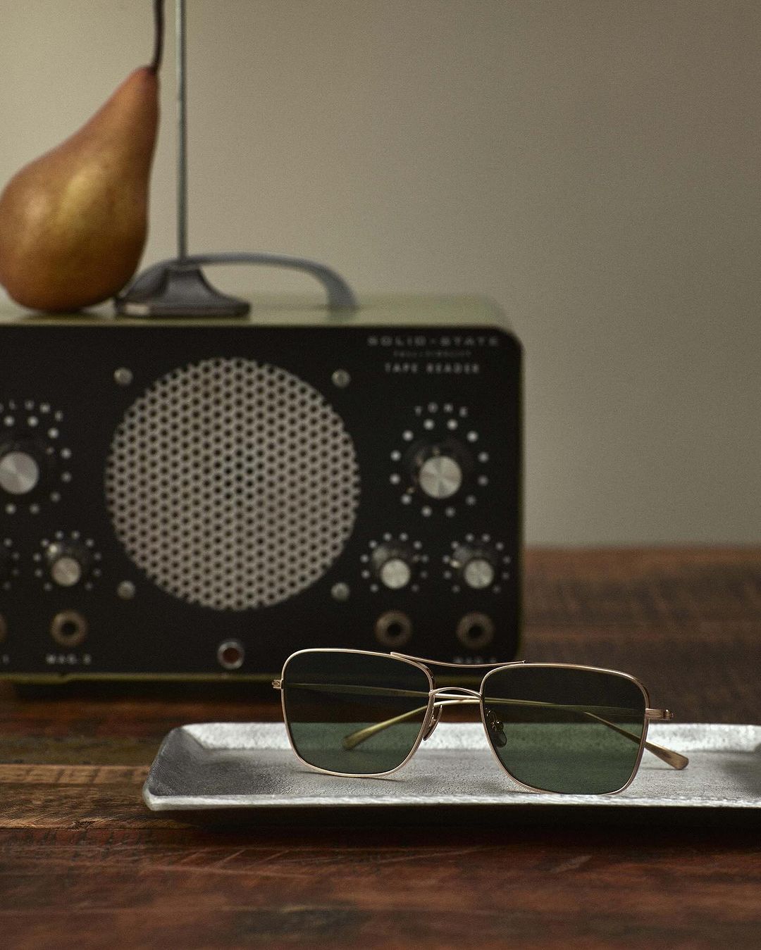 Sunglasses sit on a silver tray, with a pear and a vintage radio in the background on a wooden table against a neutral wall.
