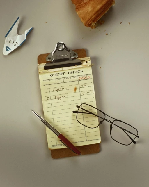 Overhead shot of a restaurant guest check on a clipboard with a pen, glasses, a torn ticket, and part of a croissant. 