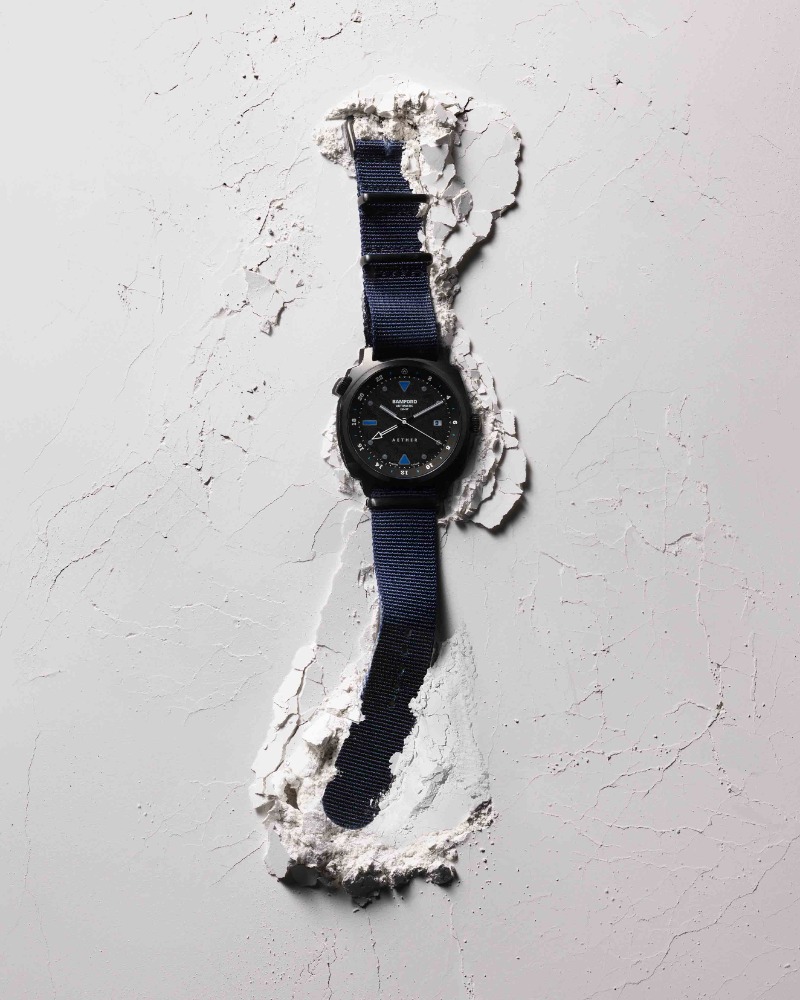 Wristwatch with a black case and blue nylon strap displayed in a stylized fashion, surrounded by textured white material for product promotion.