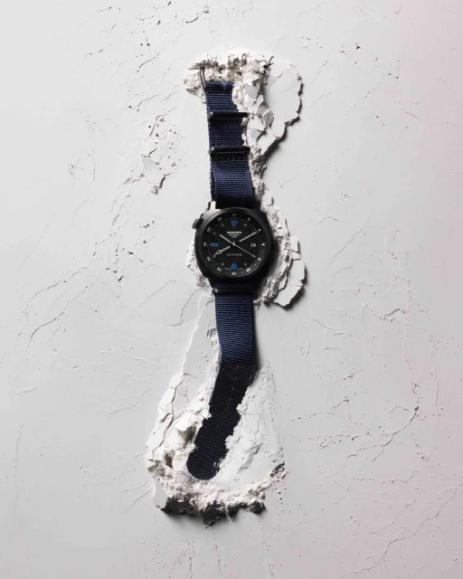 Wristwatch with a black case and blue nylon strap displayed in a stylized fashion, surrounded by textured white material for product promotion.