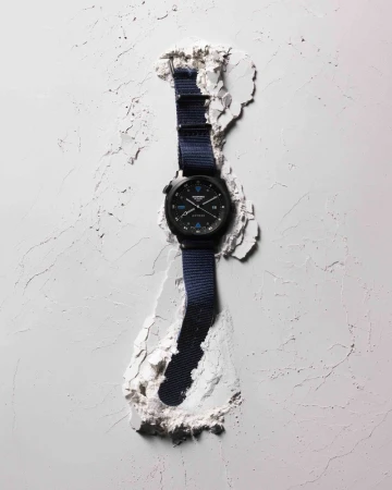 Wristwatch with a black case and blue nylon strap displayed in a stylized fashion, surrounded by textured white material for product promotion.