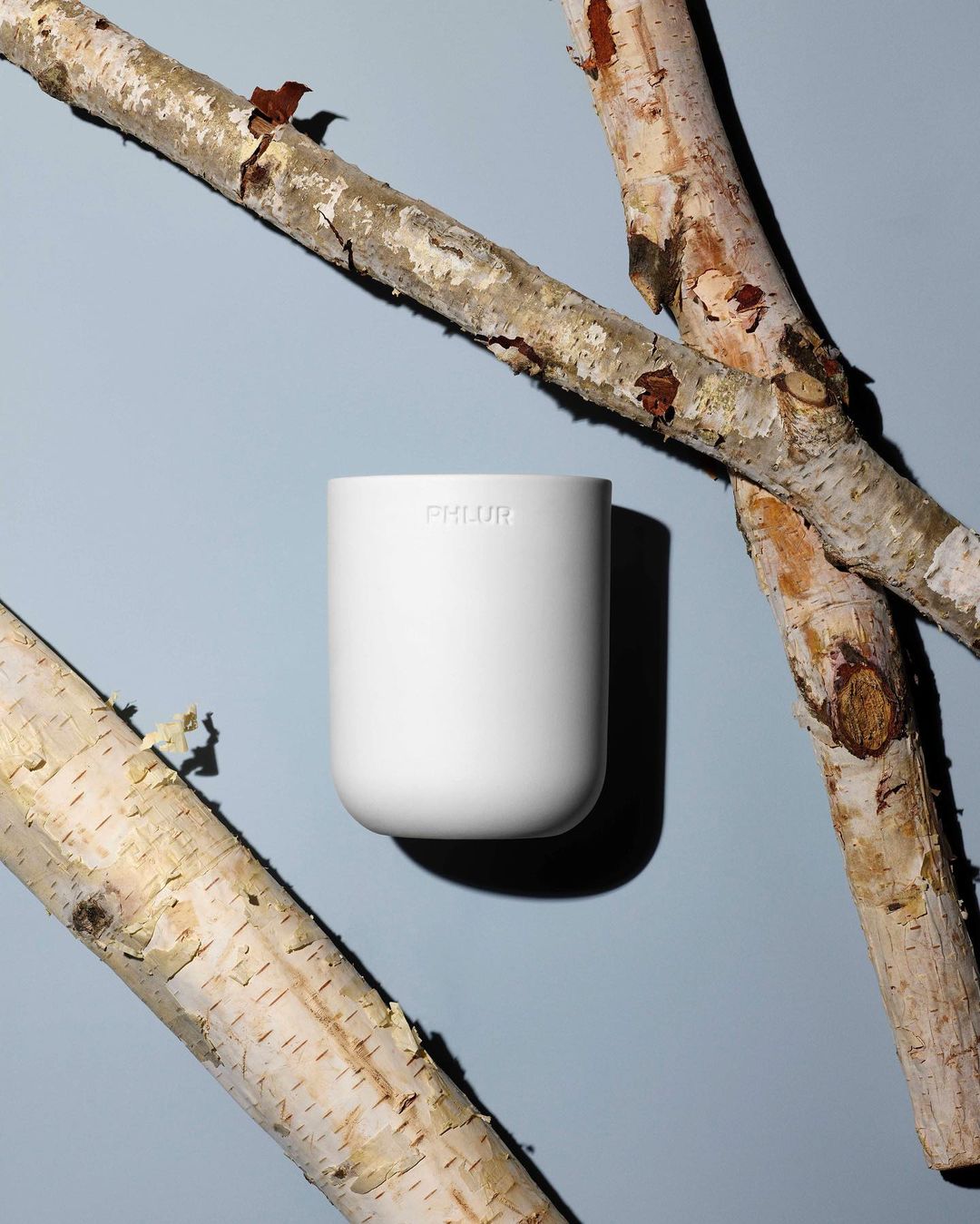 Minimalist studio shot of a white PHLUR candle in a white holder, diagonally framed by birch branches on a light blue backdrop.