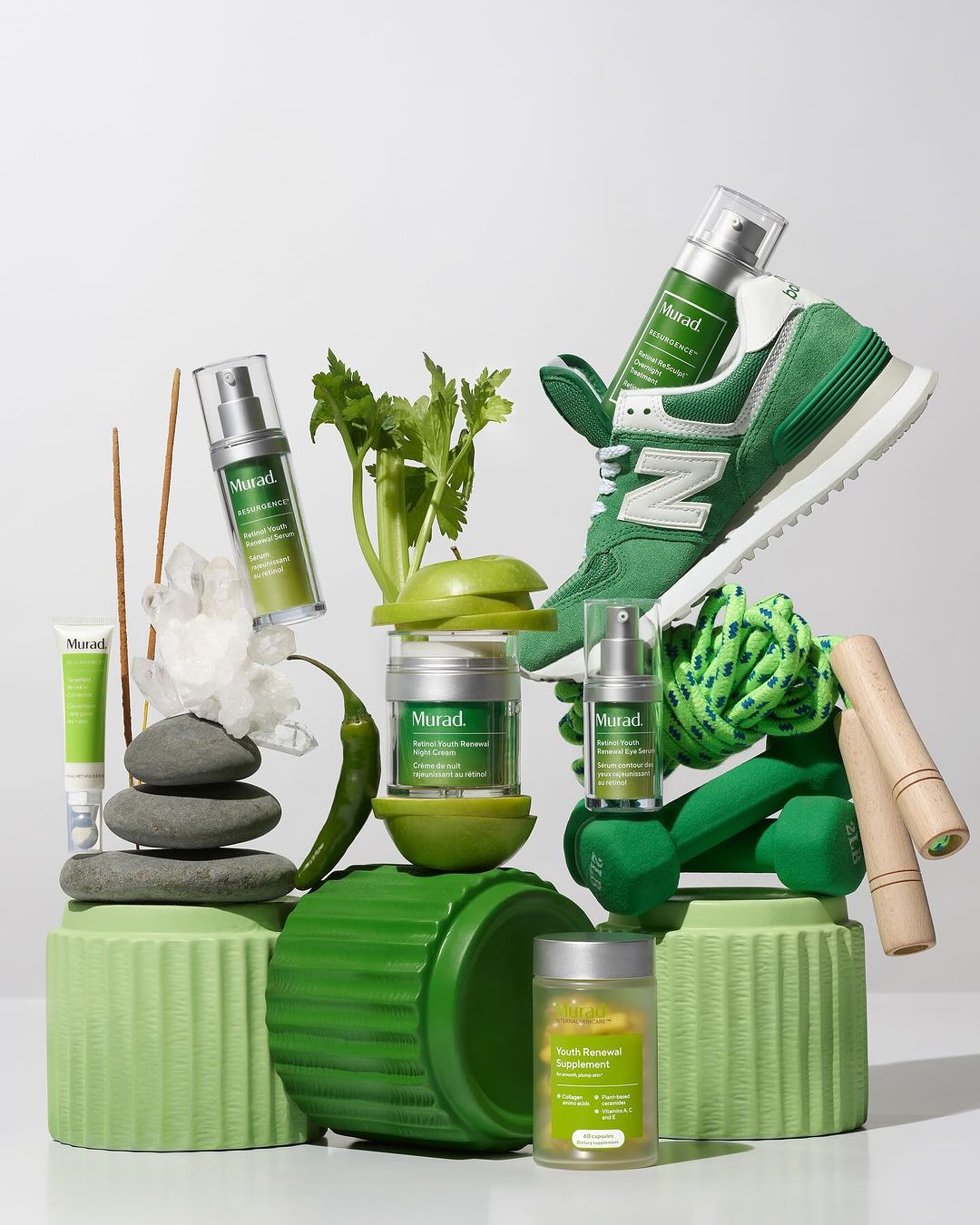 Still life featuring Murad skincare products with exercise equipment and produce, all arranged on green pedestals against a white background.