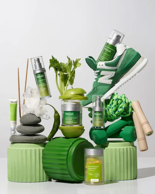Still life featuring Murad skincare products with exercise equipment and produce, all arranged on green pedestals against a white background.
