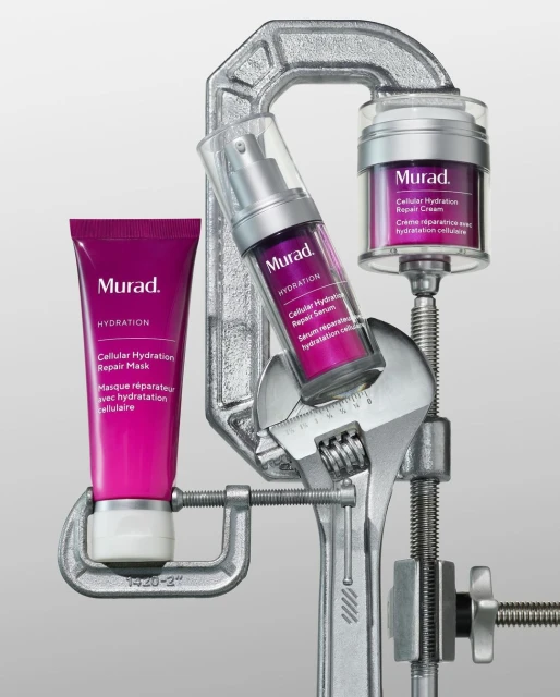 Three Murad skin care products are suspended in a silver metal clamp and wrench against a gradient gray background in a studio shot.