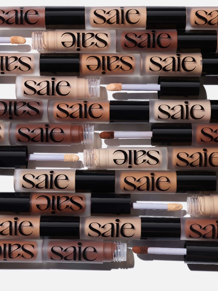 Multiple 'Saie' brand concealer tubes arranged in rows on a white surface, showcasing a variety of skin tones and product packaging.