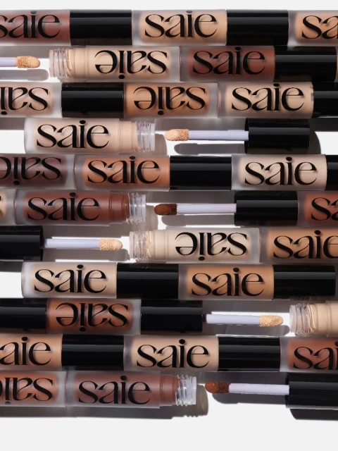 Multiple 'Saie' brand concealer tubes arranged in rows on a white surface, showcasing a variety of skin tones and product packaging.