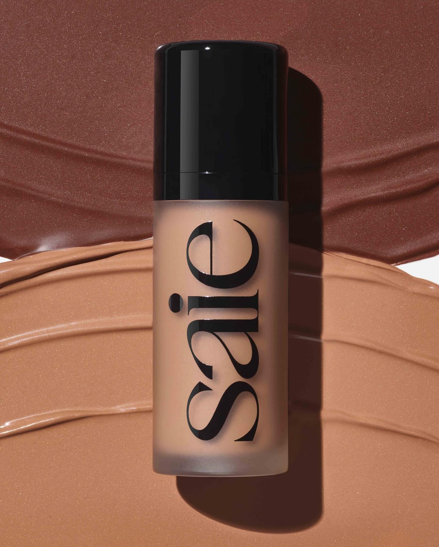 Saie brand bottle of foundation in a vertical product shot, surrounded by smears of foundation in similar warm brown shades.