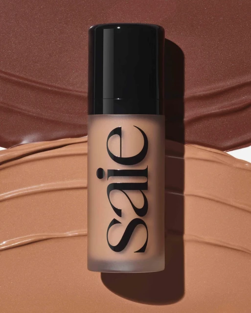 Saie brand bottle of foundation in a vertical product shot, surrounded by smears of foundation in similar warm brown shades.