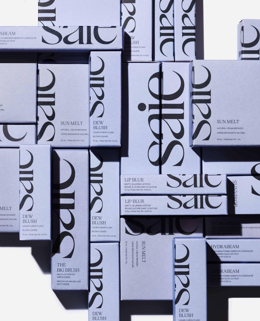 Multiple packages of Saie beauty products are arranged on a surface, including blush, bronzer, and concealer, with a pale lavender background.