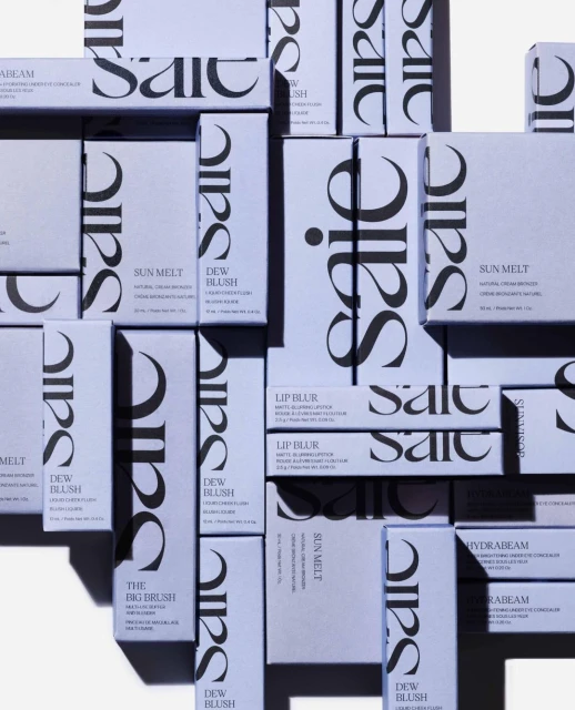 Multiple packages of Saie beauty products are arranged on a surface, including blush, bronzer, and concealer, with a pale lavender background.