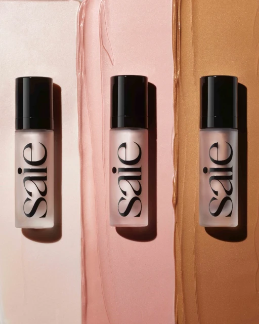 Three Saie brand cosmetic bottles lined up on a background of pale pink, rosy pink, and tan makeup smear, studio lit product shot.