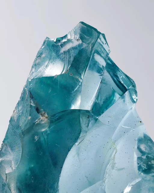 Close-up of a jagged chunk of transparent teal glass, showcasing textures and reflections against a soft, neutral background.