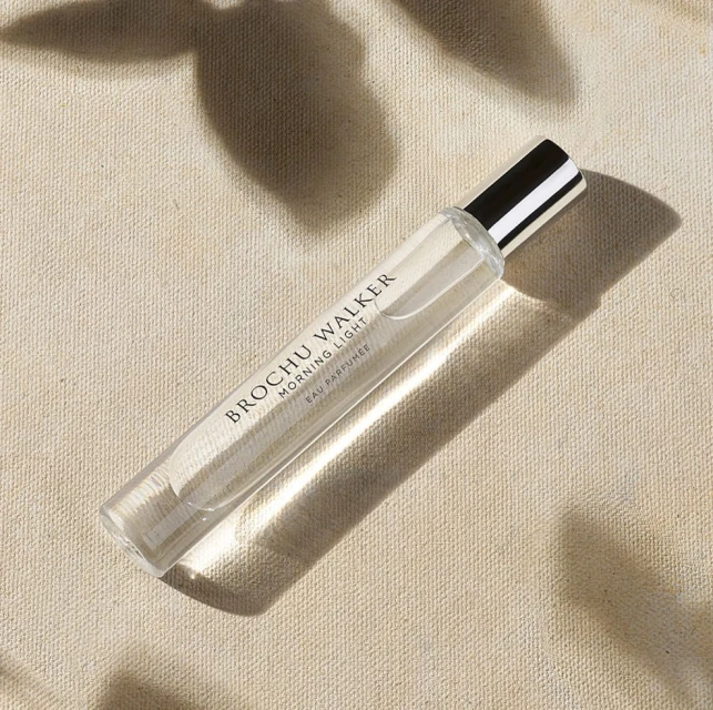 Close-up of a clear glass perfume bottle labeled BROCHU WALKER on a textured beige fabric surface with soft shadows from foliage above.