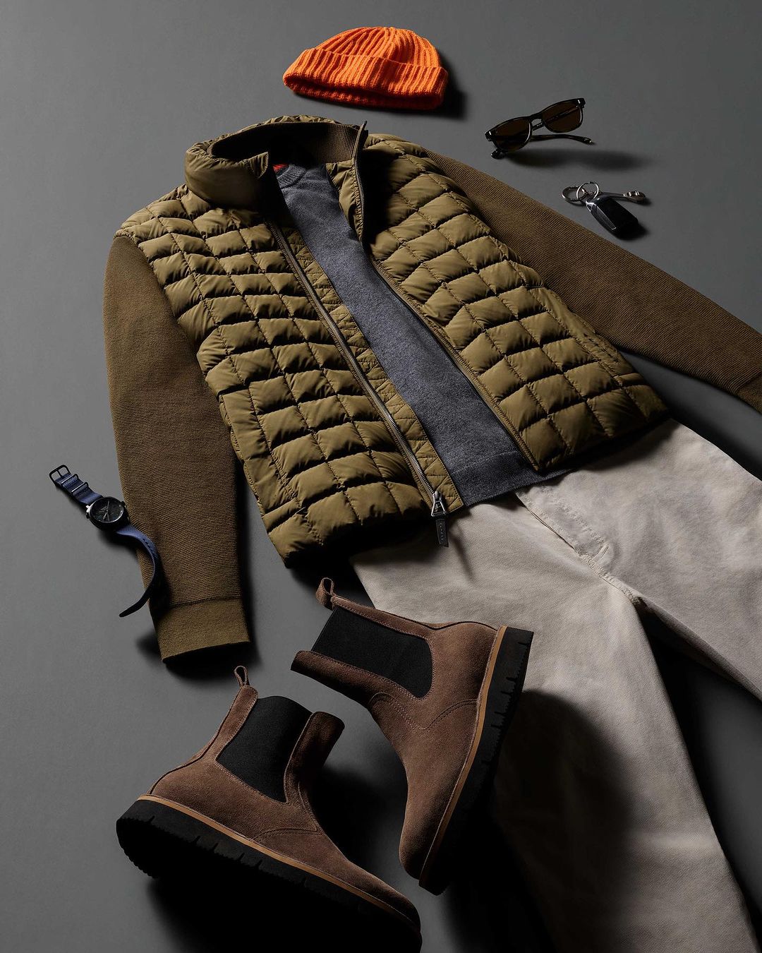 Flat lay of men's fashion apparel: olive green puffer vest, gray sweater, beige pants, brown boots, orange beanie, and accessories on gray.